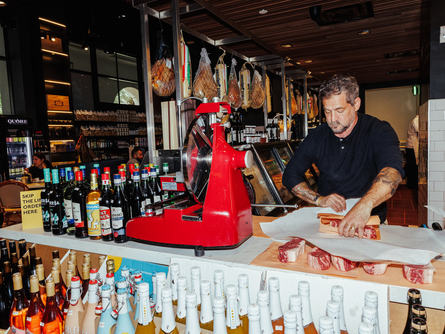 Man working at a bar with various bottles and a red meat slicer.