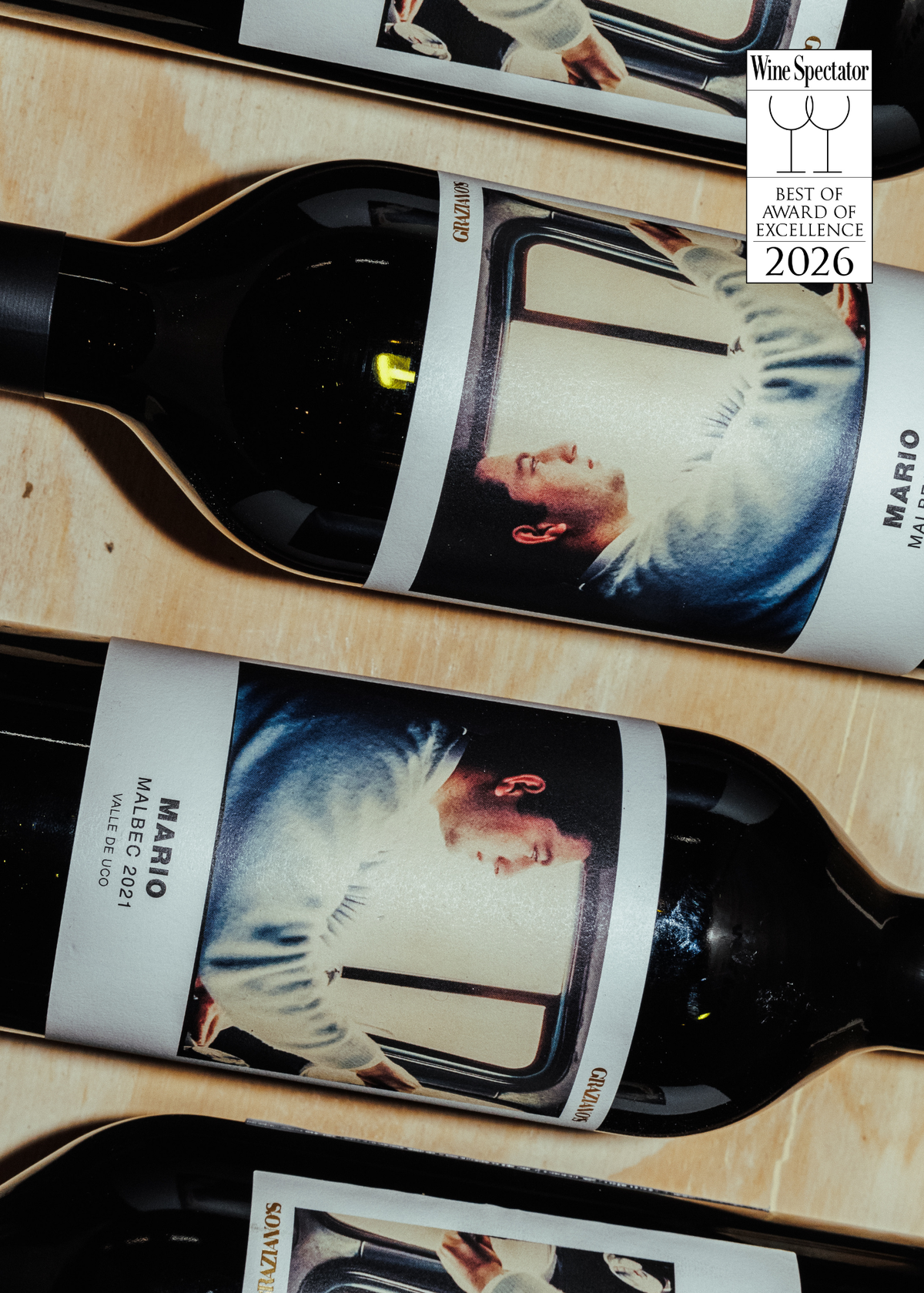Three dark glass wine bottles with custom white labels featuring a portrait of Mario, arranged diagonally in a wooden box. A Wine Spectator 'Best of Brands of Excellence 2026' badge is visible in the top right corner.