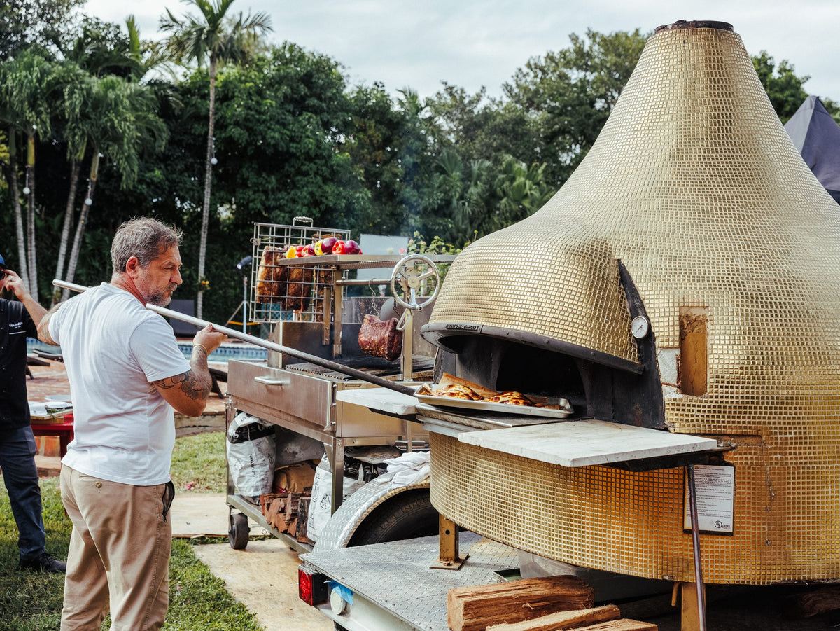 Pulling a pizza from the Graziano's wood-fired oven next to the asador at a catered event
