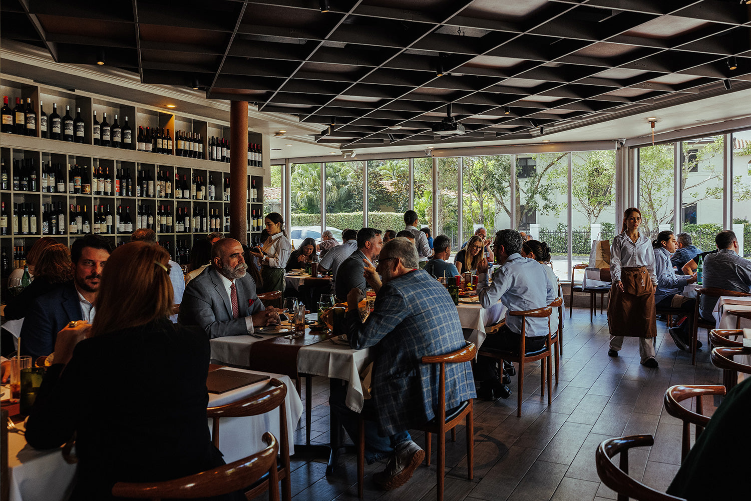 Dining area with people seated at tables, large window wall, and wine bottles on shelves.