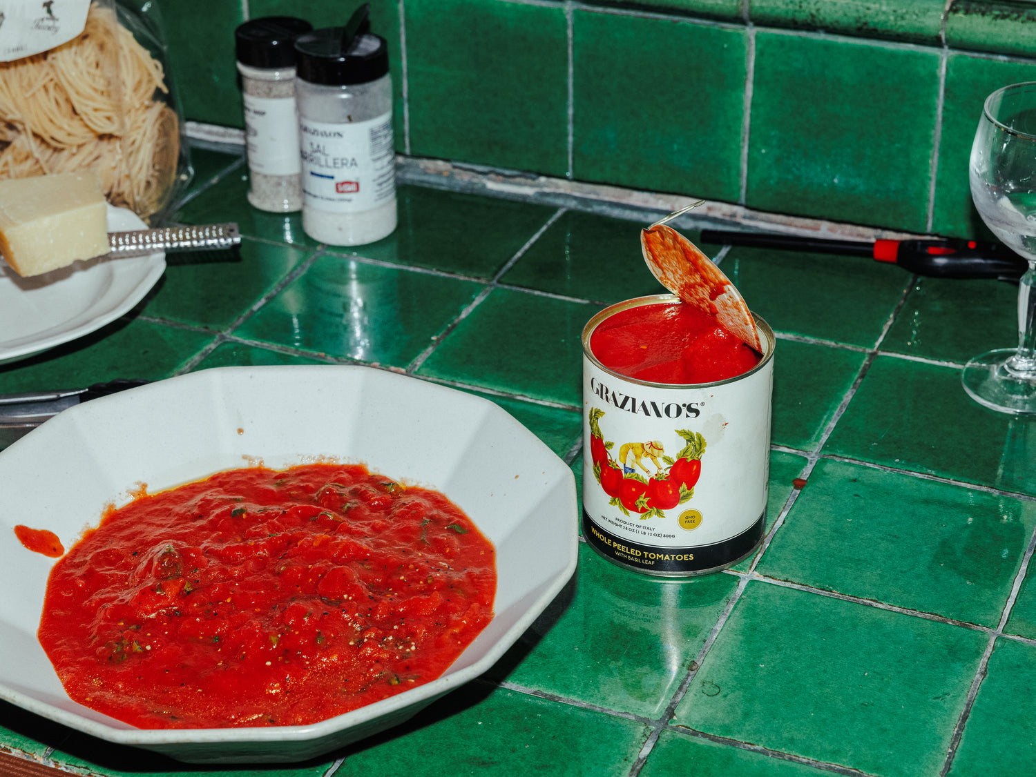Can of Graziano's tomato sauce with a spoonful of sauce on a plate and can lid, against a green tiled wall.